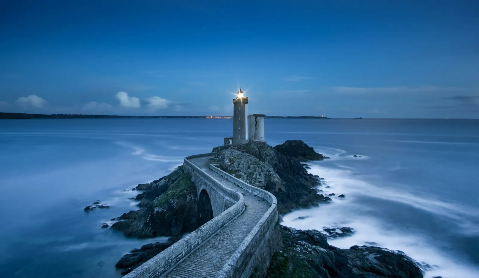 Lighthouse shining at dusk at the end of a winding stone causeway